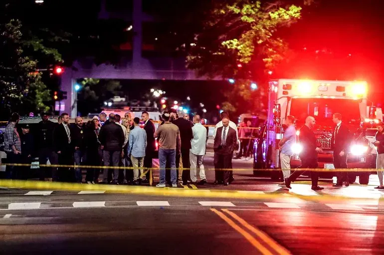 حادثة أمنية في مدينة بعد إطلاق نار، حيث يتجمع رجال الشرطة والمواطنون حول موقع الحادث، مع وجود سيارات إسعاف في الخلفية.