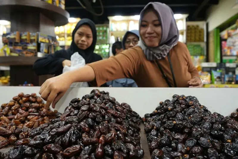 نساء يرتدين الحجاب يتسوقن في سوق، بينما تلتقط إحداهن التمر من بين كميات متنوعة، مما يعكس تقاليد الإفطار في رمضان.