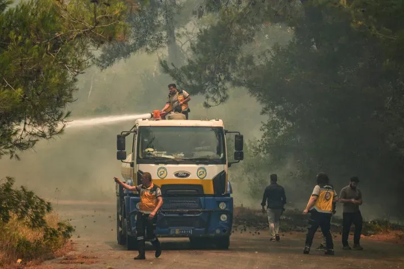رجال إطفاء يعملون على إخماد حرائق الغابات في منطقة جبل التركمان بسوريا، بينما تتصاعد الأدخنة في الخلفية.