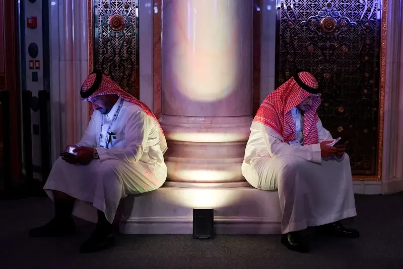 رجلان سعوديان يجلسان على الأرض بجوار عمود، مشغولان بهواتفهم المحمولة في بيئة مضاءة بشكل خافت، تعكس التوجه نحو التكنولوجيا الحديثة.