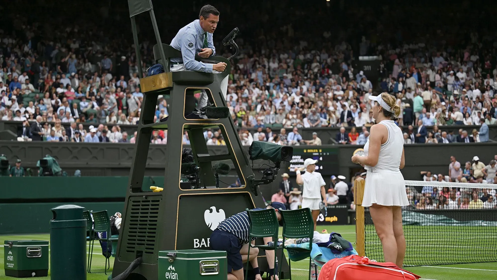 حكم مباراة تنس يجلس في كرسي مرتفع أثناء مواجهة بين لاعبتين على ملعب ويمبلدون، مع جمهور كبير في الخلفية.