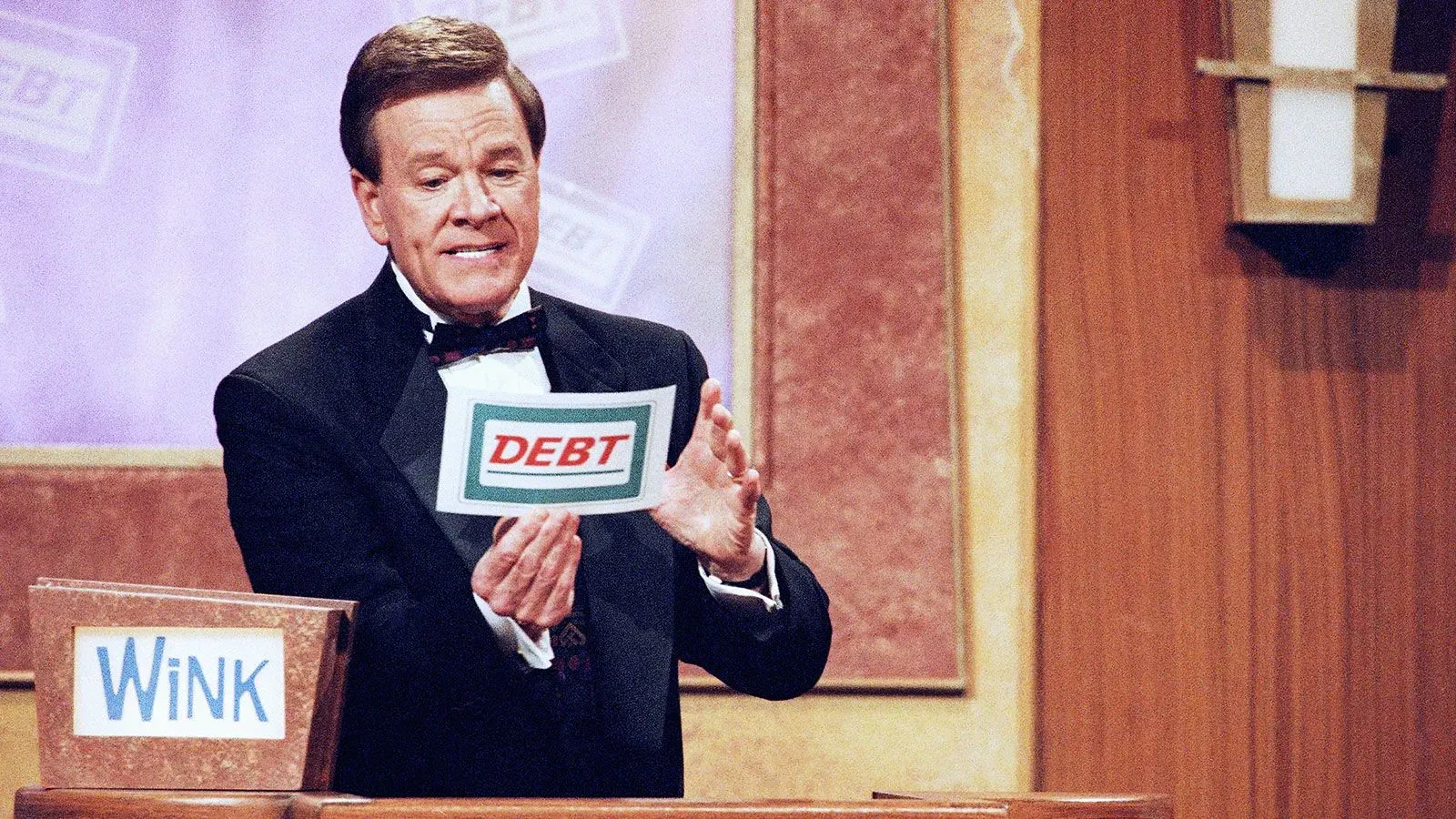 وينك مارتينديل يقدم لعبة "ديون" في برنامج تلفزيوني، مبتسمًا ويعرض بطاقة مكتوب عليها "DEBT"، مع خلفية من الاستوديو.