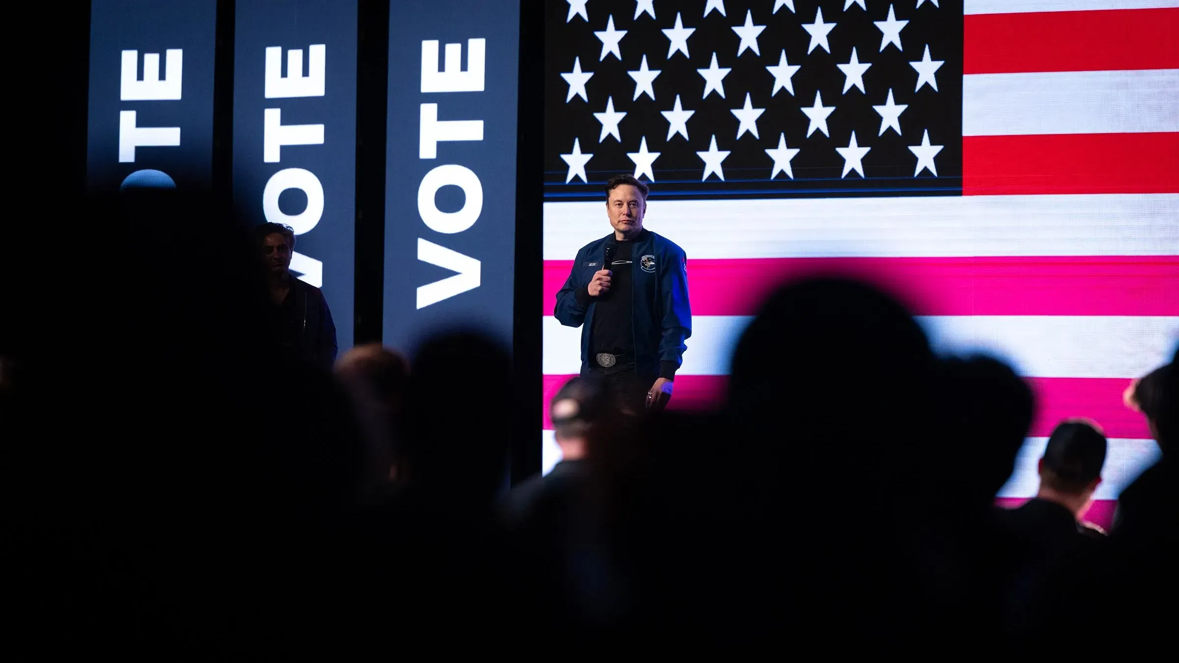 إيلون ماسك يتحدث في تجمع انتخابي لدعم المرشح الجمهوري في ويسكونسن، مع لافتات "صوت" وخلفية علم الولايات المتحدة.