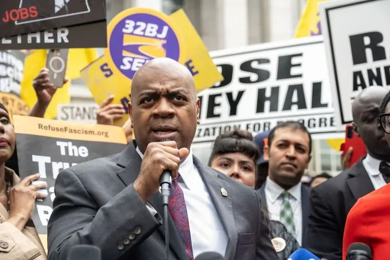 نائب يحمل ميكروفونًا ويتحدث في تجمع احتجاجي، محاطًا بأشخاص يحملون لافتات تعبر عن قضايا اجتماعية وسياسية.