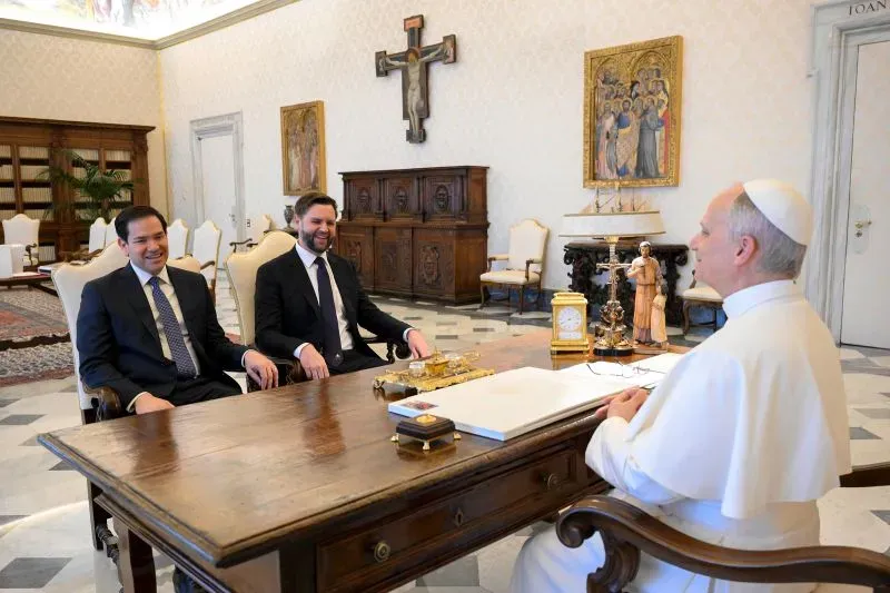 اجتماع بين شخصين يجلسان أمام البابا في قاعة مزينة، حيث يبدو النقاش غير رسمي مع ابتسامات وتعبيرات ودية.