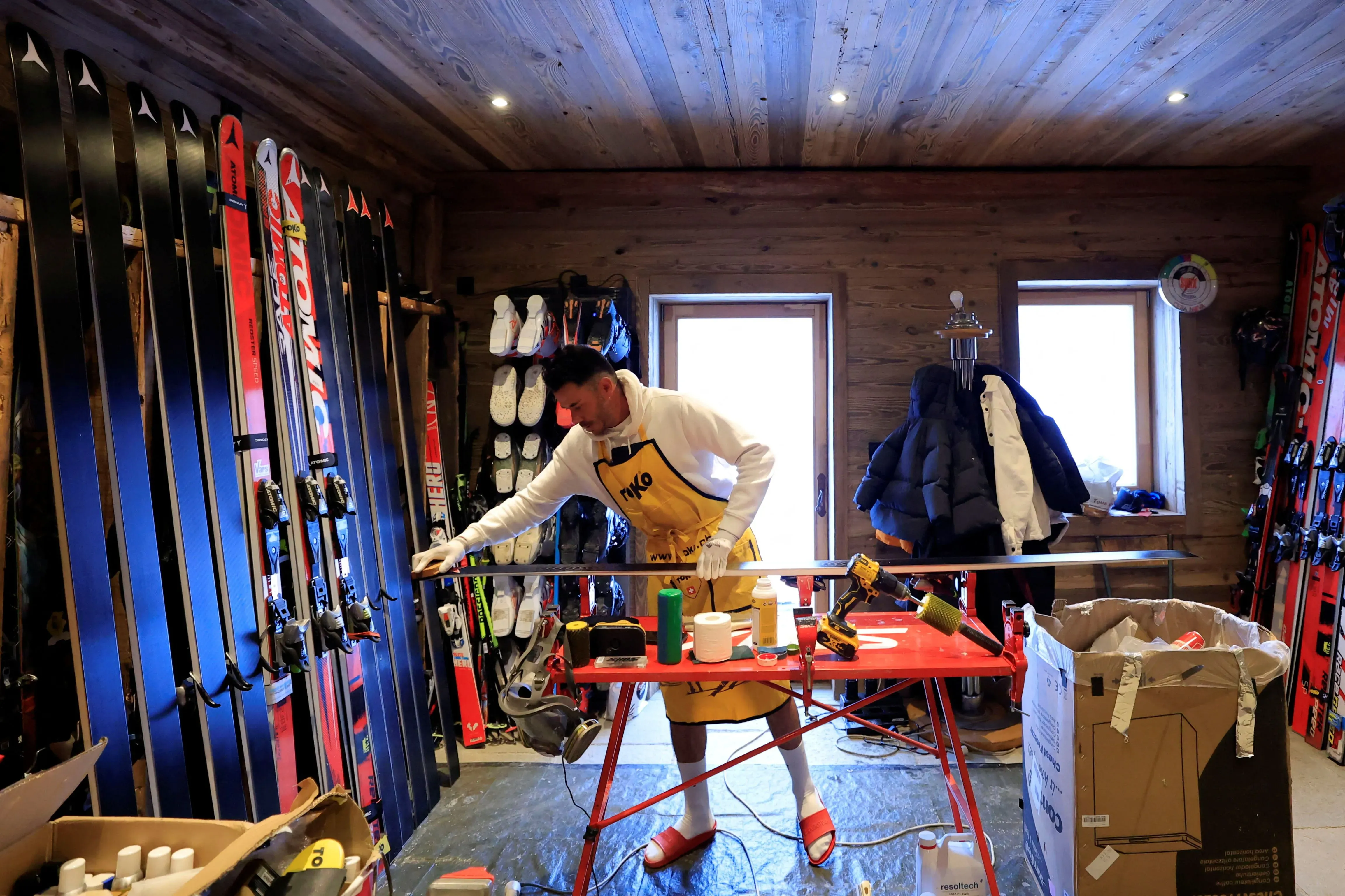 شخص يرتدي سترة صفراء ويعمل على صيانة زلاجات في ورشة خشبية، محاطًا بزلاجات أخرى وأدوات. يركز على إعداد الزلاجات للتزلج السريع.