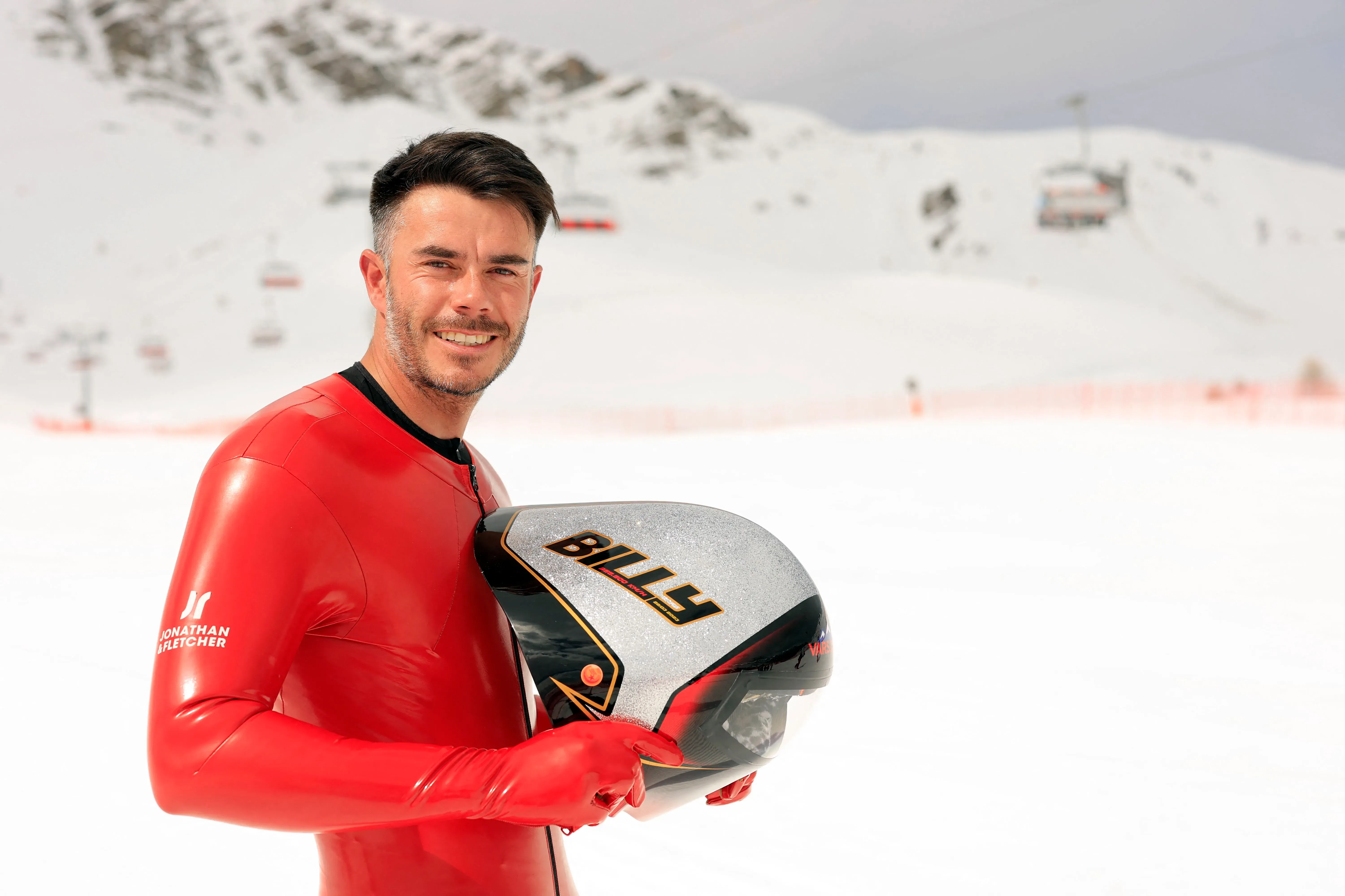 سيمون بيلي، المتزلج السريع، يرتدي بدلة تزلج حمراء زاهية وخوذة تحمل اسمه، مبتسمًا أمام المنحدرات الثلجية في فرنسا.