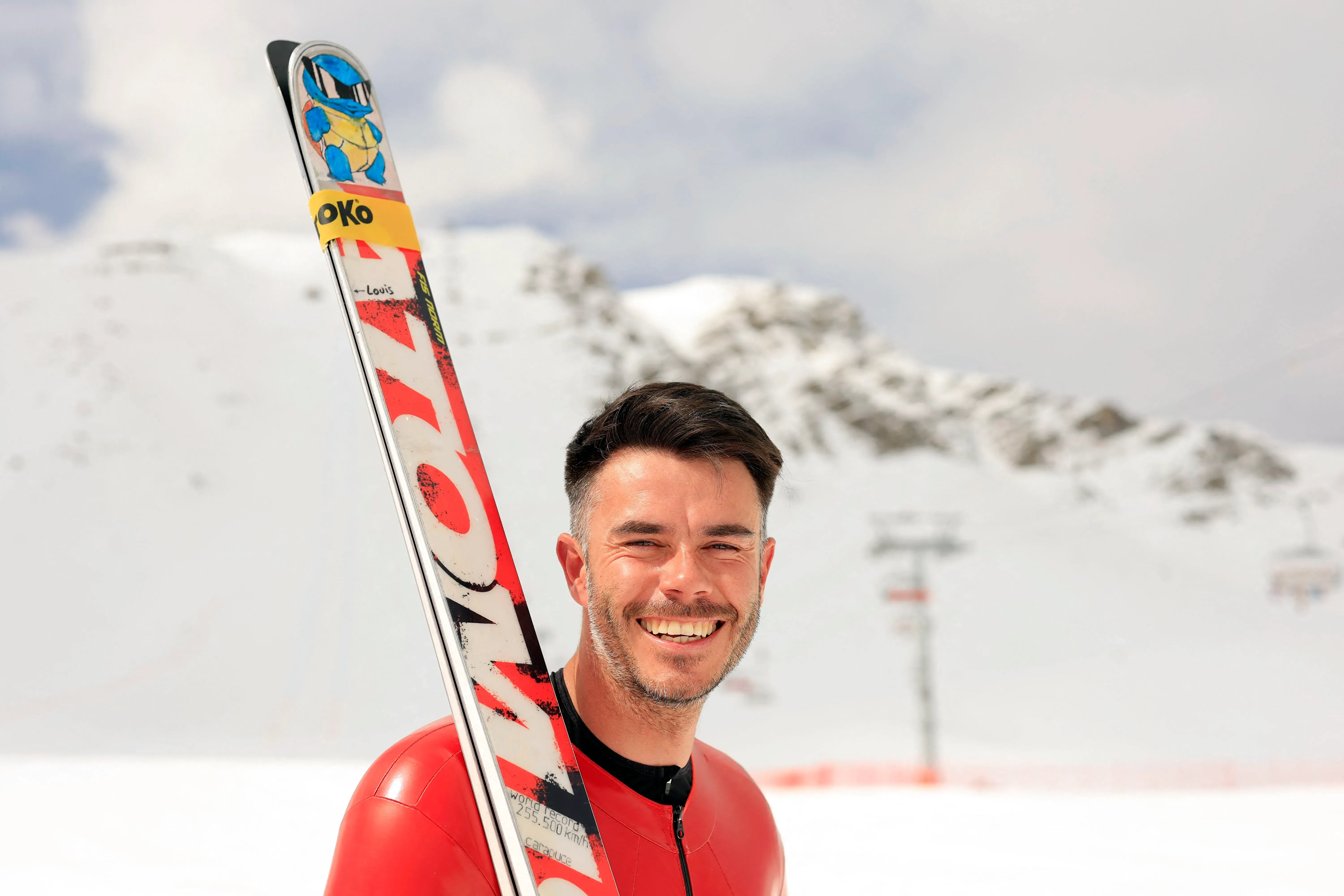 سيمون بيلي، المتزلج الفرنسي، مبتسم ويرتدي بدلة تزلج حمراء وخوذة، يقف أمام المنحدرات الثلجية، مستعدًا لتحقيق أسرع سرعة تزلج في العالم.