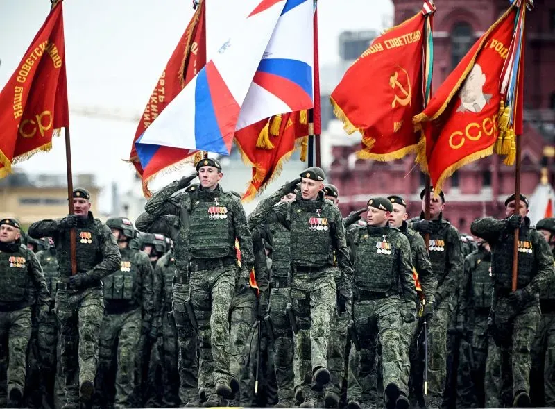 جنود روس يرتدون زيهم العسكري أثناء عرض يوم النصر في موسكو، حاملين الأعلام الروسية والسوفيتية، مع خلفية تاريخية تعكس أهمية الحدث.