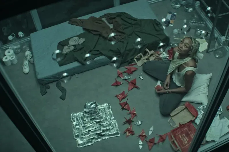 امرأة تجلس في زنزانة محاطة بأشياء شخصية، تنظر إلى السقف بعبوس، مع وجود طائرات ورقية حمراء حولها، تعكس معاناتها في مسلسل "أنت".