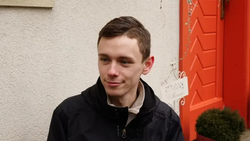 شاب يجلس أمام باب ملون، يعبر عن آرائه حول السياسة والهجرة، ويظهر اهتمامه بالحزب اليميني المتطرف في ألمانيا.