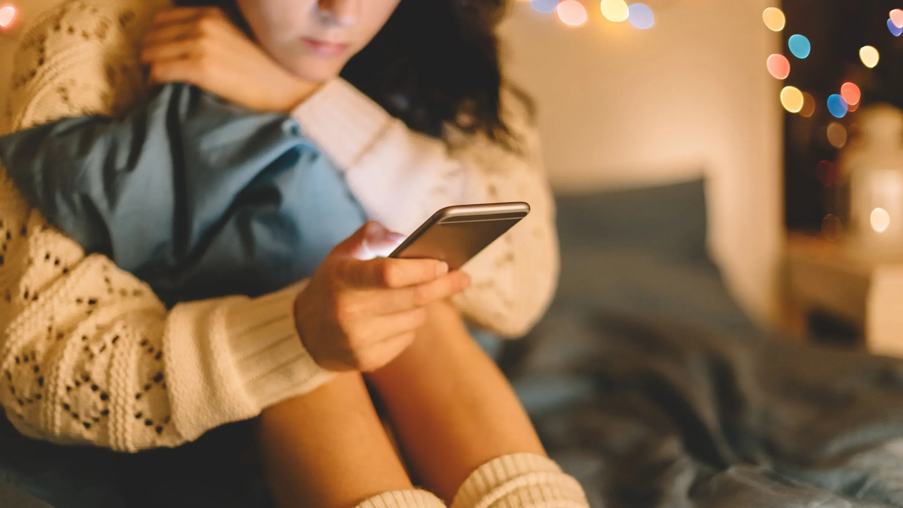 شخص يجلس في السرير ممسكًا بهاتفه الذكي، مع إضاءة خافتة وزينة ضوئية في الخلفية، تعكس عادات استخدام الشاشة وتأثيرها على الأطفال.